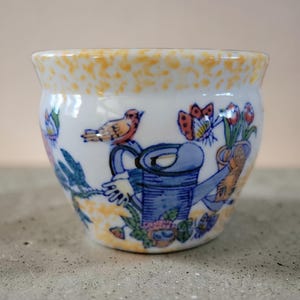 May include: A small, white ceramic planter with a yellow speckled rim. The planter features a hand-painted design of a blue watering can, a bird, a butterfly, and flowers. The design is in shades of blue, orange, and green.