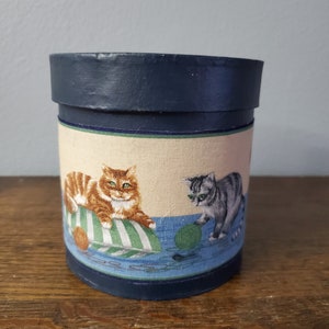 Puede incluir: Una caja redonda cubierta de tela azul y blanca con un diseño de dos gatos jugando con hilo. El gato de la izquierda es naranja y el gato de la derecha es gris.