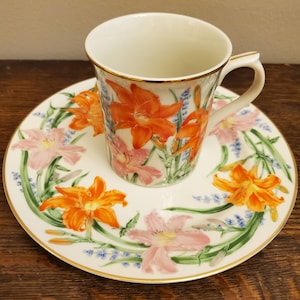 May include: A white teacup and saucer set with a floral design. The cup and saucer feature a pattern of orange and pink lilies with green leaves and blue flowers.