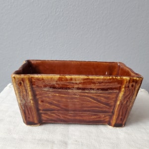 May include: A rectangular brown ceramic planter with a wood-grain texture. The planter has a slightly flared shape and sits on four small feet. The interior is a darker brown, and the exterior has a glossy finish.