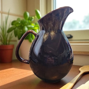 May include: A dark brown ceramic pitcher with a rounded body and a curved handle. The pitcher has a glossy finish with lighter brown accents near the spout. A wooden spoon and a plant in a terracotta pot are in the background.