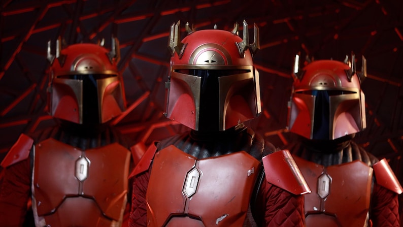 Puede incluir: Tres figuras con armadura mandaloriana roja y dorada se encuentran en un espacio con poca luz. Los cascos tienen un dise&ntilde;o distintivo con una visera en forma de T y una estructura similar a una corona. La armadura incluye petos y hombreras.