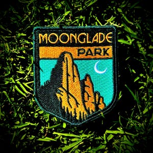 May include: An embroidered patch with a black border featuring the text MOONGLADE PARK in gold letters. The design depicts a golden mountain range against a bright turquoise sky with a white crescent moon, all rendered in detailed thread work.