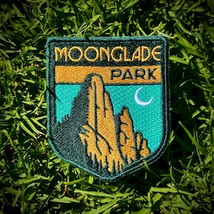 May include: A shield-shaped embroidered patch featuring the text MOONGLADE PARK in gold thread. The design depicts a dark green mountain silhouette against a teal sky with a white crescent moon. The border is dark green with gold accents.