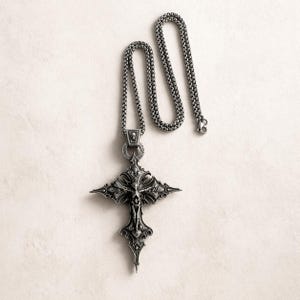 May include: A silver-toned pendant necklace featuring a detailed cross design. The cross has intricate carvings and a figure at its center. The chain is a linked style, and the pendant includes a skull detail. The necklace is displayed on a neutral background.