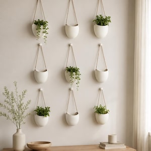 May include: Nine white, semi-circular wall-mounted planters are arranged in a grid. Each planter hangs from a thin brown cord attached to a small white knob. Several planters contain green leafy plants, while others remain empty.