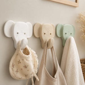May include: Three wall-mounted hooks shaped like elephant heads in white, light beige, and sage green. Each hook features a wood grain texture and a curved trunk that serves as the hook. A floral bib, a beige bag, and a white towel hang from them.