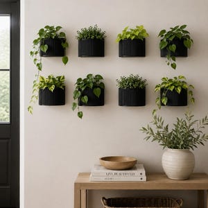 Puede incluir: Ocho maceteros de pared negros, semicirculares y acanalados, dispuestos en dos filas, cada uno con una planta verde. Debajo, una mesa consola de madera sostiene un cuenco de madera, dos libros titulados "LIVE BEAUTIFULLY" y "HOMBODY", y un jarrón.