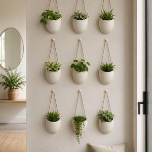 May include: A set of nine off-white, egg-shaped wall planters arranged in a three-by-three grid. Each ceramic vessel is suspended by a thin, light-colored cord attached to a small wall hook, containing a variety of green indoor plants.