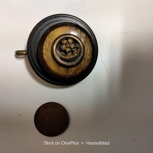 May include: A circular pendant featuring a dark brown outer ring, a lighter wood-grain center, and a small brass floral emblem in the middle. A small metal loop is attached to the side. A dark brown circular coin is placed below the pendant.