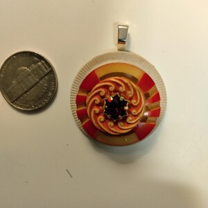 May include: A circular pendant featuring a layered design with a white ridged outer edge, a red and yellow striped middle section, and a central orange swirl pattern topped with a dark red faceted stone, shown next to a US nickel for scale.