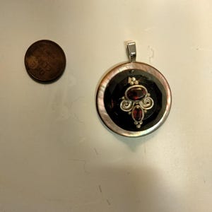 May include: A circular pendant featuring a dark, faceted center stone surrounded by an iridescent shell border. A silver-toned metal setting holds two oval red stones and decorative scrollwork. A copper penny is shown for scale.