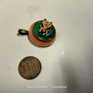 May include: A circular pendant featuring a green button base topped with a small gold-toned bee ornament with clear crystals, positioned next to a copper one-cent coin for scale. The pendant has a bronze-colored metal loop for attachment.
