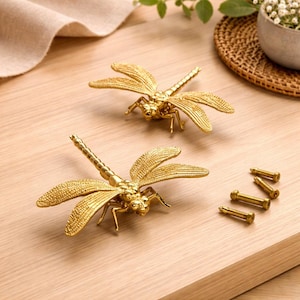May include: Two gold-colored dragonfly cabinet pulls with detailed wings and bodies. The pulls are displayed on a light wood surface, along with several gold-colored screws. Decorative hardware for furniture.