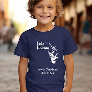 Pu&ograve; includere: Una maglietta blu navy con una grafica bianca del lago Norman, Carolina del Nord. Il testo "Lake Norman" &egrave; sopra il contorno del lago e "North Carolina's -Inland Sea-" sotto. La maglietta &egrave; indossata da un bambino.