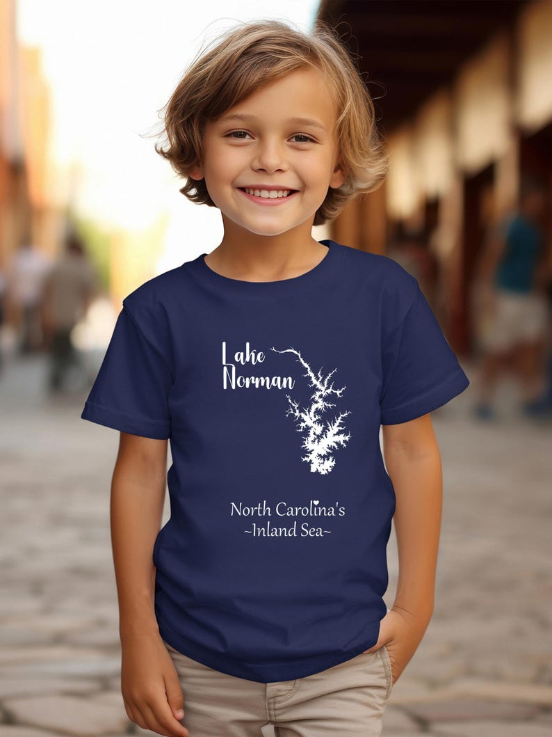 Pu&ograve; includere: Una maglietta blu navy con una grafica bianca del lago Norman, Carolina del Nord. Il testo "Lake Norman" &egrave; sopra il contorno del lago e "North Carolina's -Inland Sea-" sotto. La maglietta &egrave; indossata da un bambino.