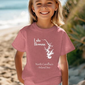 Pu&ograve; includere: Una maglietta rosa polveroso con la scritta "Lake Norman" e un disegno bianco del lago. Sotto c'&egrave; la scritta "North Carolina's - Inland Sea-". La maglietta &egrave; indossata da una persona giovane che sorride alla fotocamera.