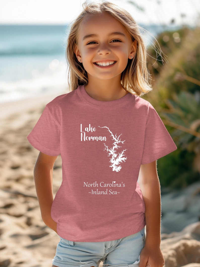 Pu&ograve; includere: Una maglietta rosa polveroso con la scritta "Lake Norman" e un disegno bianco del lago. Sotto c'&egrave; la scritta "North Carolina's - Inland Sea-". La maglietta &egrave; indossata da una persona giovane che sorride alla fotocamera.