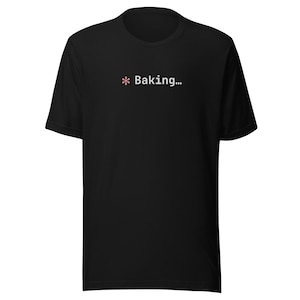 Può includere: T-shirt nero con la scritta "* Baking..." in bianco. L'asterisco è un fiore rosa. La maglietta è a girocollo e a maniche corte.