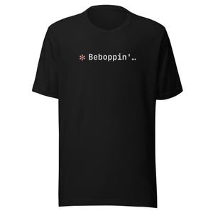 Può includere: T-shirt nero con la scritta "* Beboppin'..." in bianco, con un asterisco rosa. La maglietta è a girocollo e maniche corte.