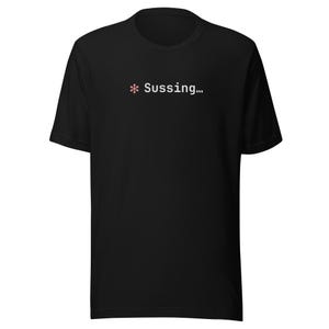Può includere: T-shirt nero con la scritta "* Sussing..." in bianco e rosa. La maglietta ha un girocollo ed è realizzata in un tessuto morbido.