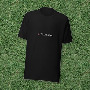 Puede incluir: Una camiseta negra de manga corta con un pequeño símbolo de asterisco rojo seguido del texto blanco "Thinking..." centrado en el área del pecho. La prenda tiene un cuello redondo estándar y un dobladillo recto, con un diseño sencillo.