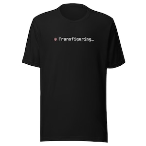 Può includere: T-shirt nero con la scritta "* Transfiguring..." in bianco e rosa. La maglietta girocollo sembra essere realizzata in un materiale morbido. Il testo è centrato sul petto.