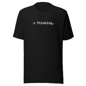 Può includere: T-shirt nera con la scritta bianca "Thinking...", preceduta da un piccolo simbolo floreale rosa. La maglietta ha maniche corte e girocollo.