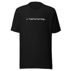 Könnte beinhalten: Schwarzes T-Shirt mit dem Text "* Tomfoolering..." in Weiß, mit einem kleinen rosa Sternchen. Das T-Shirt mit Rundhalsausschnitt ist ein schlichtes Design, das für den Alltag geeignet ist. Der Text ist mittig platziert.