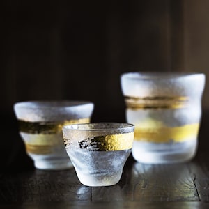 May include: Three frosted glass tumblers with gold bands. The glasses vary in size and are arranged on a dark wooden surface. The gold bands have a textured, brushed appearance, adding a touch of elegance to the glassware.