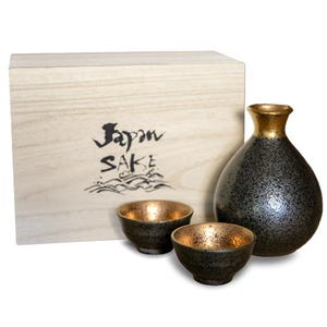 May include: A Japanese sake set comprising a wooden box with "Japan Sake" text, a black and gold sake bottle, and two matching cups. The sake bottle and cups have a speckled design with gold interiors.
