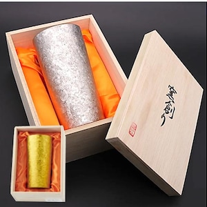 May include: A silver-coloured cylindrical drinking vessel with a textured surface, presented in a wooden box with orange satin lining. A smaller gold-coloured vessel is in a separate box. The boxes have Japanese text.