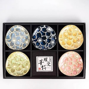 May include: A set of six decorative ceramic plates in a black box. Each plate features a floral design in shades of blue, yellow, pink, and green. The plates are arranged in a grid pattern, with a small label in the center.