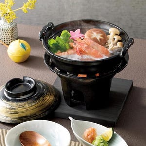 Pode incluir: Uma panela quente japonesa preta com alças, cheia de camarões, cogumelos e vegetais. A panela fica em um suporte preto com um pequeno queimador. Uma tampa com detalhes dourados e pratos de servir estão por perto. Um recipiente amarelo em forma de limão e um vaso com flores amarelas também estão presentes.