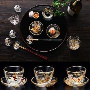 Puede incluir: Un conjunto de tazas de sake de cristal transparente y platos pequeños con bordes dorados e ilustraciones de gatos pintadas. El set incluye una bandeja circular negra grande, una botella y dos pares de palillos, con tazas y platillos adicionales debajo.