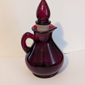 May include: A deep red glass cruet with a rounded body, a curved handle, and a pointed glass stopper. The vessel features a small pouring spout at the top rim. The glass is translucent and uniform in color, with a smooth, polished surface finish.