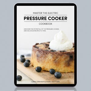 May include: A digital tablet shows a cookbook cover titled "Master the Electric Pressure Cooker Cookbook." The image features a blueberry dessert with whipped cream and fresh blueberries on a wooden board. Authors: Cami Graham & Marci Buttars.