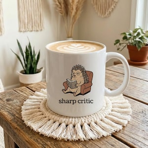 May include: White ceramic mug featuring a cartoon hedgehog reading a book, seated in a brown armchair. The mug has the words "sharp critic" printed below the hedgehog. The mug sits on a cream-coloured macrame coaster.