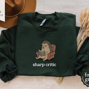 May include: Dark green sweatshirt featuring a cartoon hedgehog wearing glasses and reading a book, seated in a brown armchair. The text "sharp critic" is printed below the hedgehog. The sweatshirt has a crew neck and long sleeves.