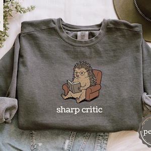 May include: Dark gray sweatshirt with a cartoon hedgehog wearing glasses and reading a book while seated in a brown armchair. The text "sharp critic" is printed below the illustration. The sweatshirt has long sleeves.