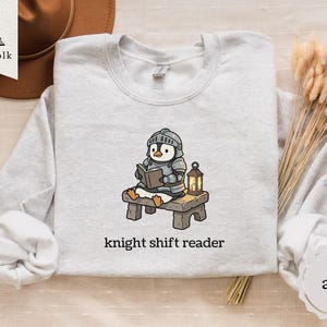 Peut inclure: Sweat-shirt gris clair avec un dessin animé de pingouin en armure de chevalier lisant un livre. Le pingouin est assis sur un petit banc en bois à côté d'une lanterne. Le texte "knight shift reader" est imprimé en dessous.