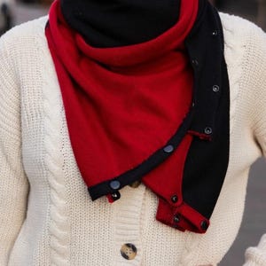 May include: A red and black scarf with button fastenings. The scarf is worn over a cream-coloured cable knit cardigan. The scarf is triangular, with the black section along the edges and the red in the centre.