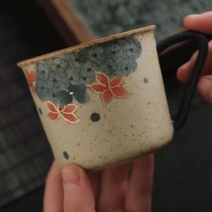 May include: A ceramic mug with a speckled beige finish, decorated with hand-painted red and blue floral designs. The mug has a black handle and a slightly flared rim, ideal for hot drinks.