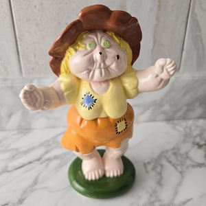 May include: A ceramic figurine of a doll-like character with a brown hat, yellow top, and orange skirt. The doll has green eyes, rosy cheeks, and a stitched patch on the clothing. It stands on a green circular base.