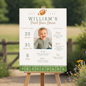 May include: A rectangular milestone sign featuring a football theme with the text William's First Year Down. It includes a central photo of a baby, player stats listing 23 pounds and 31 inches tall, and a list of favorites like songs, books, and toys.