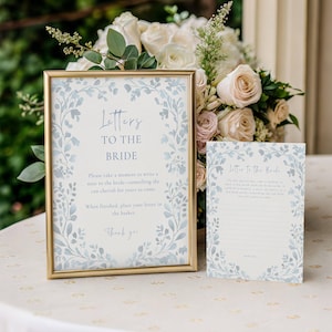 Op de afbeelding: Een goud ingelijst bord en een bijpassende kaart met blauwe aquarel bladranden en de tekst Letters to the Bride. Het bord bevat instructies om een briefje voor de bruid te schrijven om jarenlang te koesteren en een dankjewel.