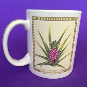 May include: White ceramic mug with a gold rim and handle. The mug features a colorful illustration of an ornamental pineapple with green leaves and pink fruit. The text "Ornamental Pineapple" is printed below the image.
