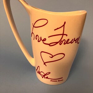 May include: White ceramic mug with a unique curved handle and a flared rim. The mug features the handwritten words "Love Forever" in burgundy script, along with a heart symbol and the signature "Donna Karan".