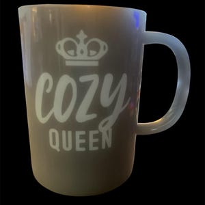 May include: A gray mug with a white handle and the words "COZY QUEEN" in a script font, topped with a crown graphic. The mug is cylindrical and appears to be made of ceramic or a similar material. The background is black.
