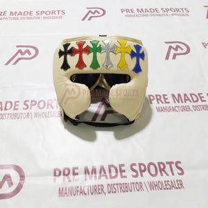 Puede incluir: Un casco de boxeo de color crema con seis parches decorativos en forma de cruz en negro, rojo, verde, plateado, amarillo y azul en la frente. El equipo de protección tiene una abertura central para los ojos y una correa para la barbilla.
